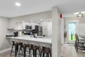 A modern kitchen with white cabinets, a built-in microwave, and a coffee bar. There are four bar stools and a dining table with chairs.