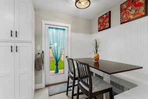 A dining area with a table, chairs, a vase with a plant, a door with a curtain, and a wall with a framed picture.