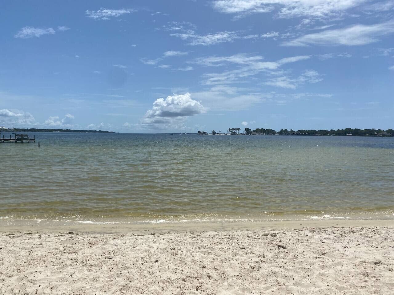 Une plage à l’eau claire, avec un rivage sablonneux et une côte lointaine parsemée de constructions.