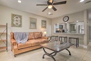 A living room with a leather couch, a coffee table, a ceiling fan, and a kitchen area with a microwave and refrigerator.
