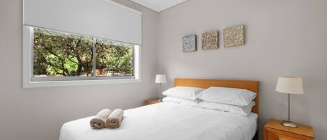 Un dormitorio con un ventanal grande, una cama con ropa blanca, dos mesitas de noche con lámparas y tres obras de arte en la pared.