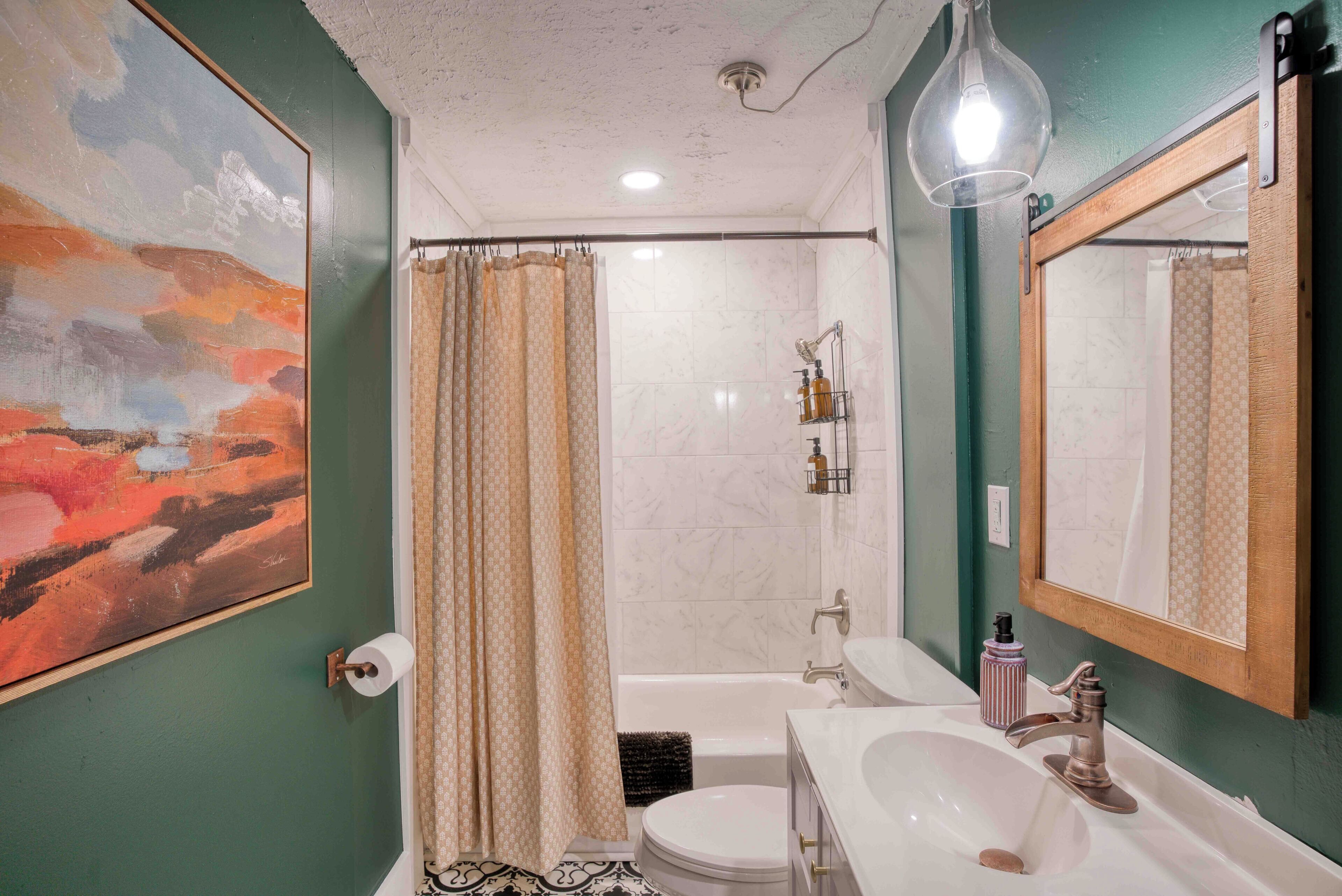 Un baño con inodoro, lavamanos, bañera, cortina de ducha, lámpara colgante y un cuadro grande en la pared.