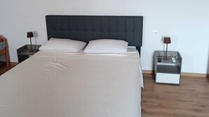 Una cama bien tendida con un cabecero oscuro, dos mesitas de noche con lámparas, y una pared lisa con un pequeño marco de fotos.