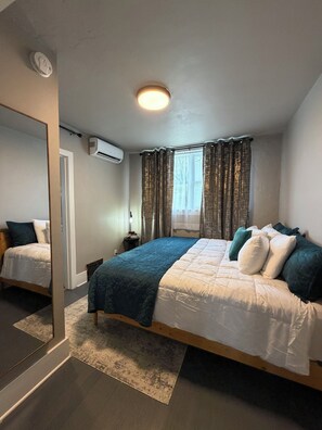 A bedroom with a bed, a bedside table, a mirror, an air conditioning unit, and a window with curtains.