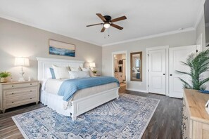 Un dormitorio con cama, lámparas de noche, ventilador de techo, alfombra estampada y una puerta que da a otra habitación.