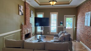 A living room with a brick fireplace, a flat-screen TV, a sectional sofa, and a door with a window. - Free Parking, Walking Distance, Camden Yards, M&T Stadium, U of MD, Convention (Baltimore)