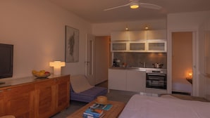 Ett kompakt bostadsutrymme med tv, matbord, soffa, ett litet kök och en takfläkt.