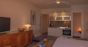 Een compacte woonruimte met een televisie, een eettafel, een bank, een kitchenette en een plafondventilator.