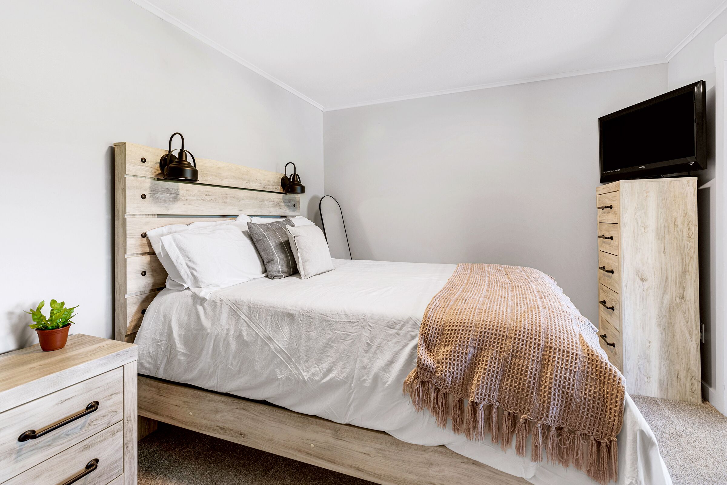 A bedroom with a wooden bed, white bedding, a television on the wall, and a small plant on the nightstand.