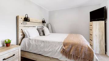 A bedroom with a wooden bed, white bedding, a television on the wall, and a small plant on the nightstand.