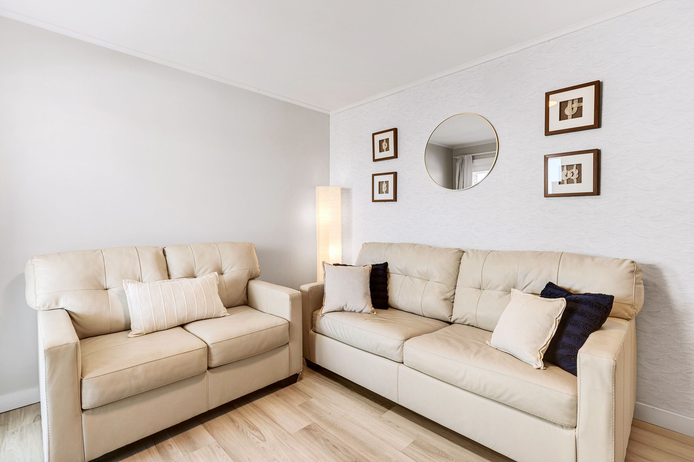 Deux canapés beiges avec des coussins, un miroir rond et des tableaux encadrés sur un mur blanc à texture.
