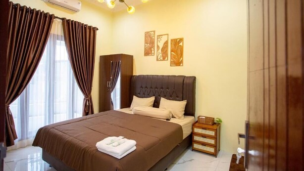 A bedroom with a large bed, curtains, a ceiling light fixture, wall art, and a wooden cabinet. - By The Padi - Homestay (Padang)