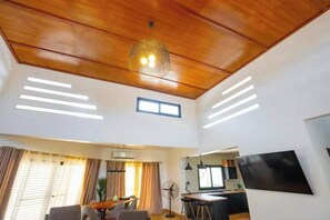 Un vaste espace de vie doté d’un plafond en bois, d’une table à manger, d’un coin cuisine et d’un grand téléviseur à écran plat fixé au mur.