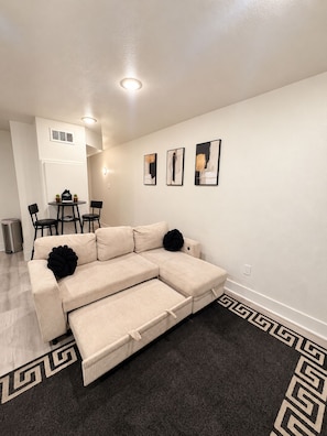 A living room with a beige sectional sofa, a dining area with a small table and chairs, and three framed artworks on the wall.