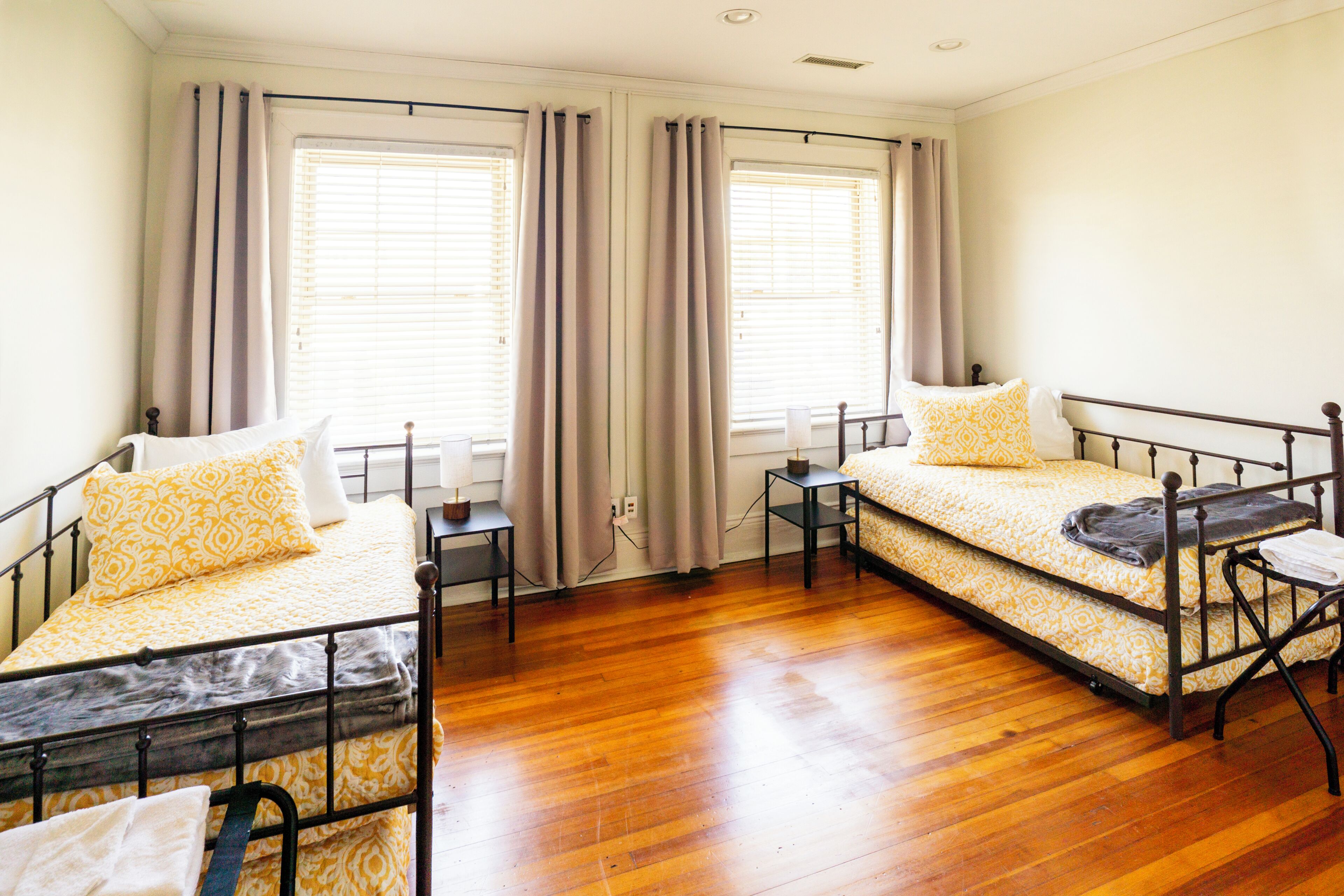 A room with two beds, wooden flooring, and large windows with curtains.