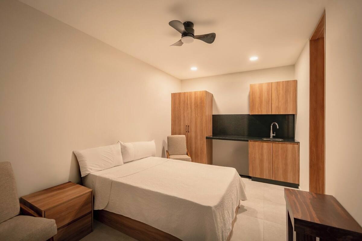 Un dormitorio con cama, mesitas de noche, una silla, ventilador de techo y una kitchenette con lavabo y armarios.
