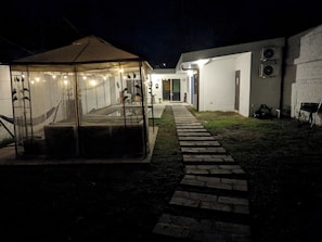 Un espace extérieur aménagé avec une zone de sièges couverte, protégée par une paroi de verre, un chemin en briques et un bâtiment à l’extérieur blanc.