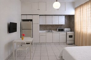 Ein kompaktes Hotelzimmer mit Küchenzeile, einem Bett, einem kleinen Tisch mit einer Vase und einem an der Wand befestigten Fernseher.