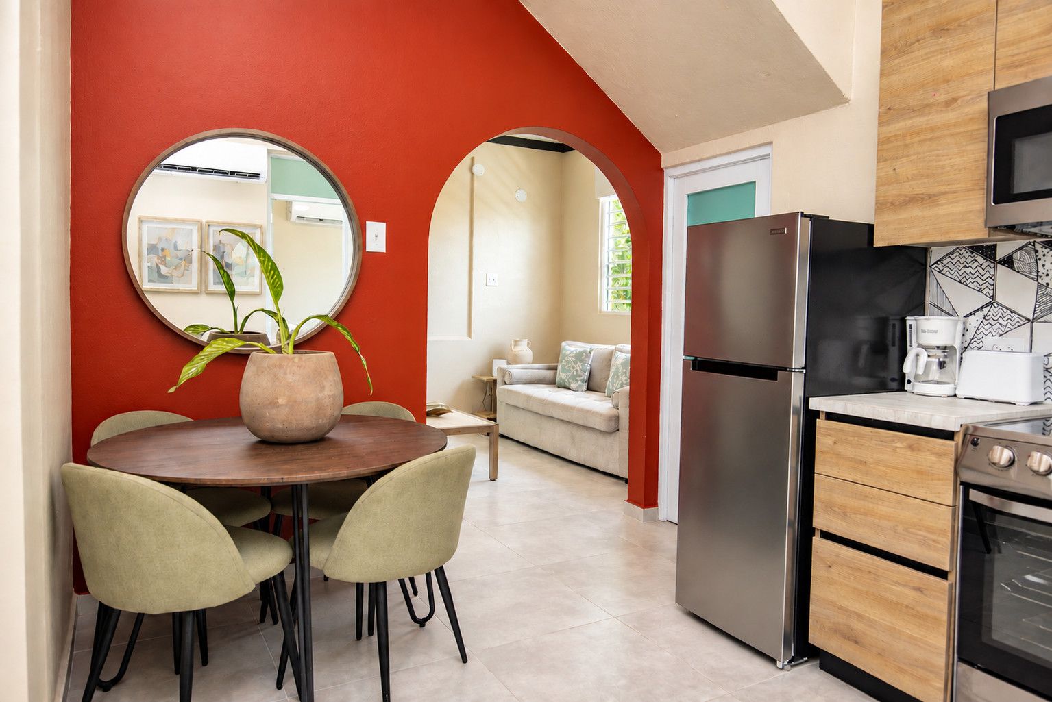 Una cocina moderna con un refrigerador de acero inoxidable, una cafetera y un microondas. Hay un comedor con una mesa redonda y sillas, además de una planta en maceta.