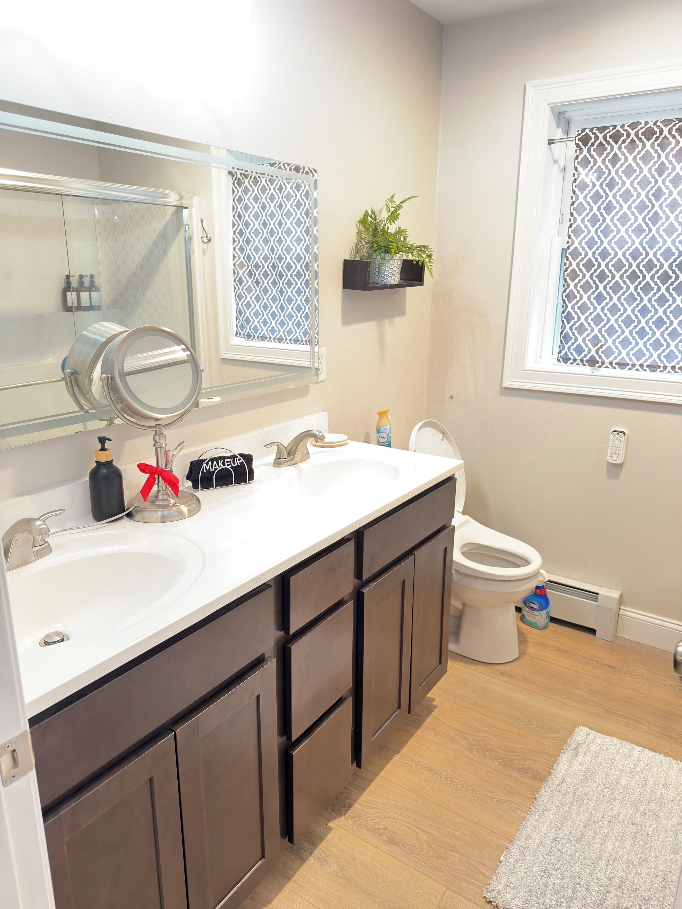 Un baño con lavamanos doble, inodoro, espejo y ventana con cortinas estampadas.