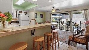 A kitchen with a bar area, wooden stools, and a view through a glass door to an outdoor seating area. - Ocean View Condo with Lanai, Pools in Kailua-Kona (Kailua Kona)