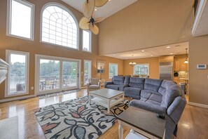 Ein geräumiges Wohnzimmer mit einem grauen Ecksofa, einem Couchtisch und einem gemusterten Teppich. Große Fenster bieten einen Blick ins Freie.
