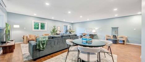 A spacious living room with a grey sofa, a round dining table with chairs, and a large window.