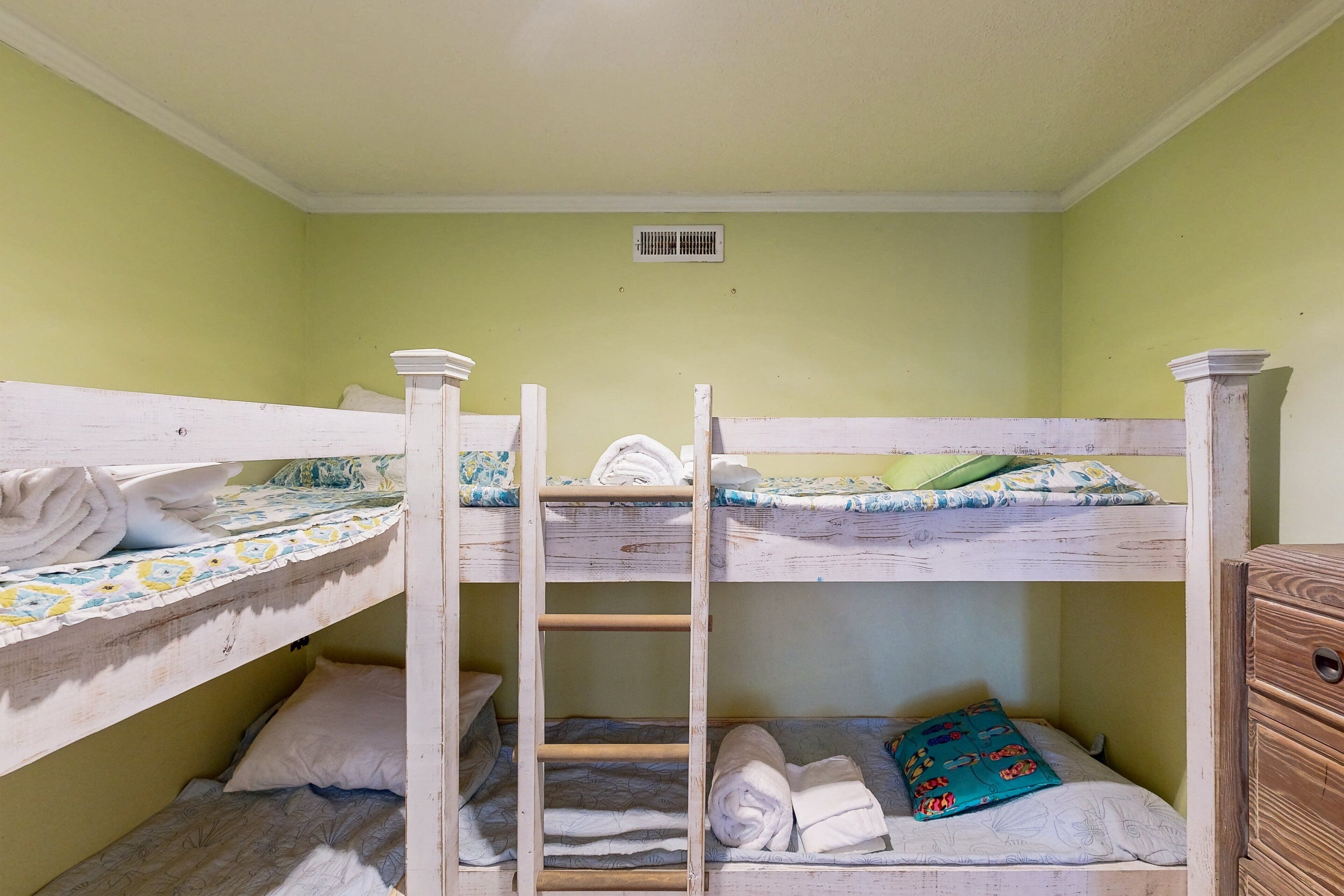 A bunk bed room with a wooden ladder, a small wooden dresser, and a vent on the wall.