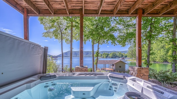 Un jacuzzi con pérgola de madera, con vista a un lago rodeado de árboles y un edificio distante.