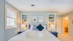 Una cama bien hecha con toallas enrolladas, una mesita de noche con una lámpara y una estantería con libros y objetos decorativos.