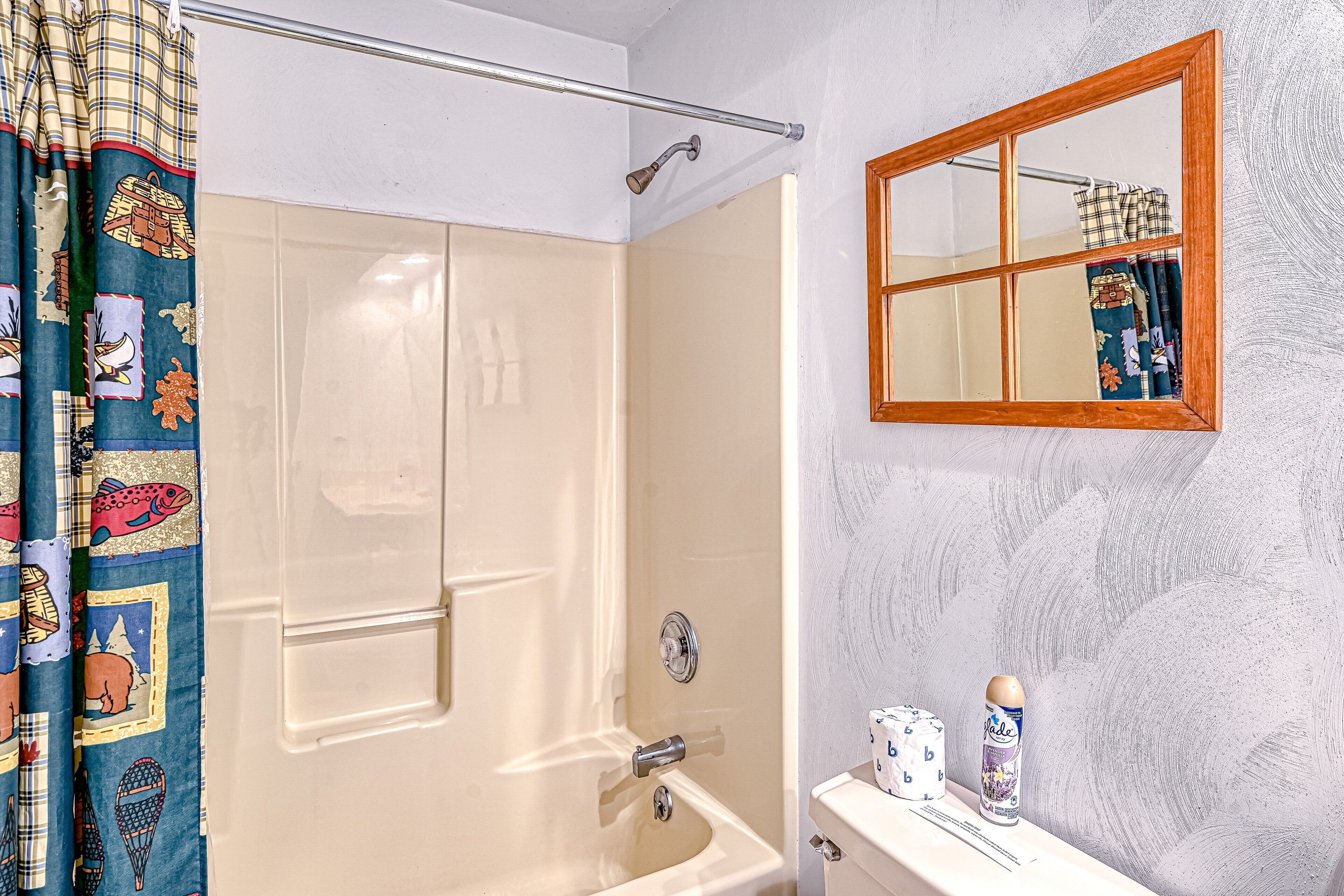 Un baño con bañera, cortina de ducha con estampado de peces, espejo con marco de madera y un inodoro con una caja de pañuelos.