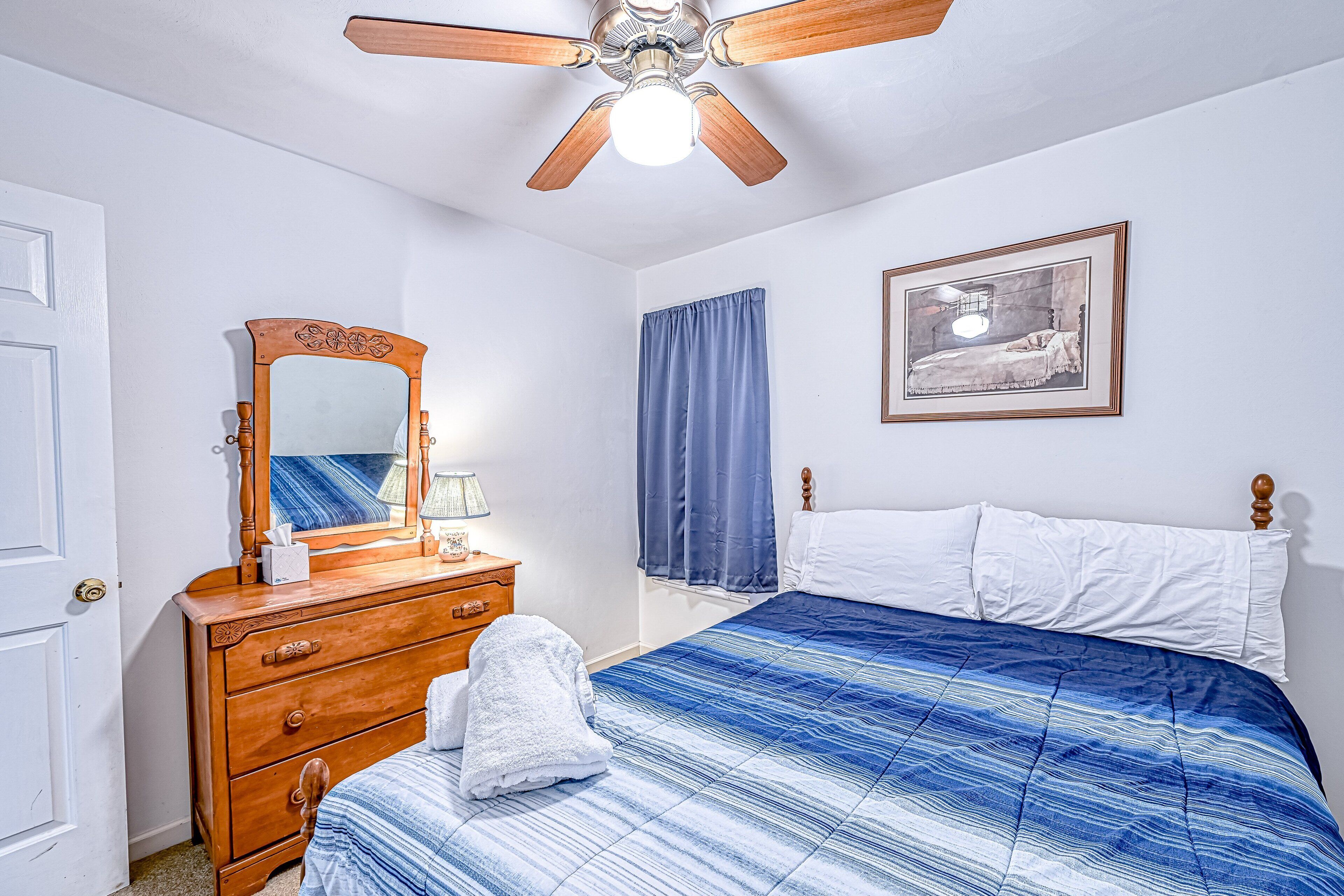 A bedroom with a wooden dresser, a bed with a blue and white striped bedspread, a ceiling fan, a framed picture on the wall, and a window with blue curtains.
