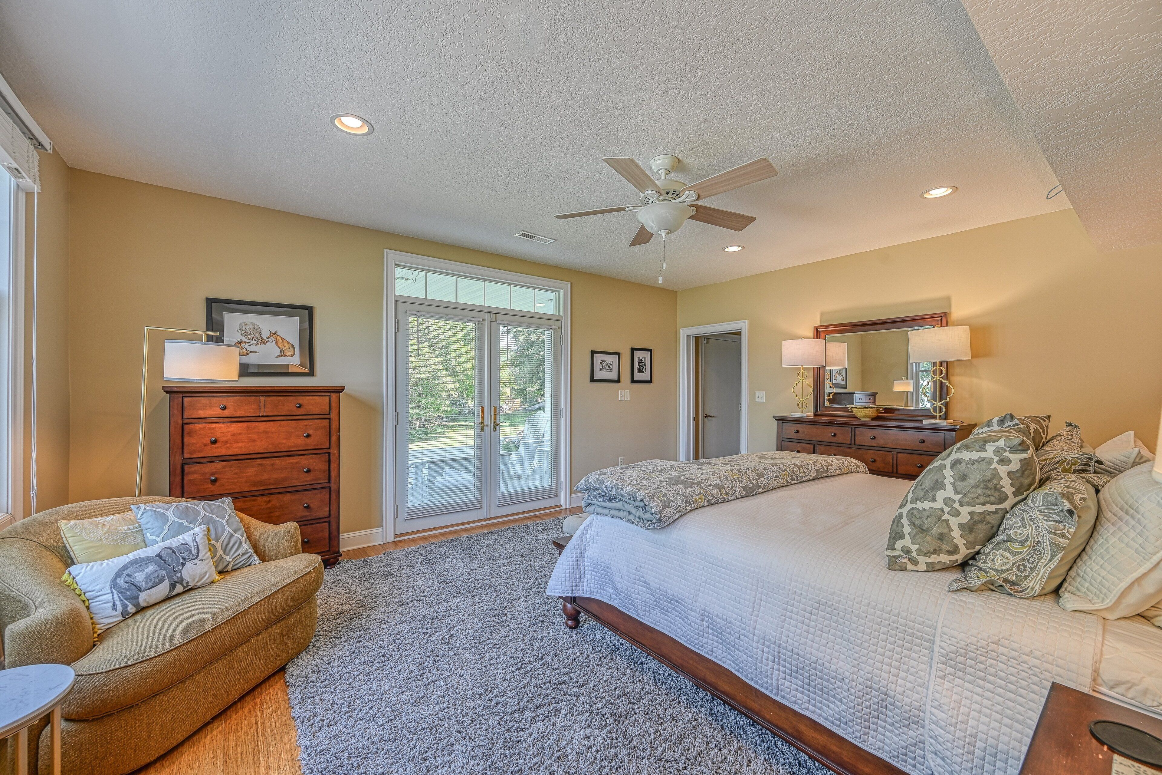 A bedroom with a bed, a dresser, a chair, a ceiling fan, and a large window.