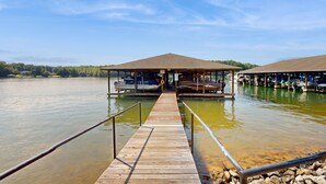 Eine Holzsteg, der zu einem überdachten Bootsanleger führt, mit freiem Blick auf das Wasser und Bäume in der Ferne.