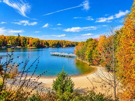 Un lago con un muelle, rodeado de árboles de colores otoñales y un cielo despejado.