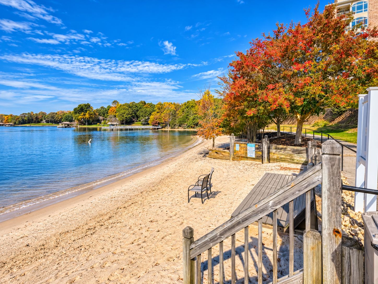 Une plage de sable avec une promenade en bois menant à un bâtiment, un lac paisible et des arbres aux feuilles d’automne.
