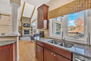 A kitchen with granite countertops, wooden cabinets, a stone fireplace, and a large window with a view of trees. - Osprey Cove - Custom Lakefront Home With 2-story Dock, Wifi, Game Room and Fireplace (Moneta)