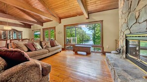 Un salon spacieux doté d’un plancher en bois, d’une cheminée en pierre et de grandes fenêtres offrant une vue sur la verdure.