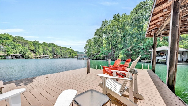 Une terrasse en bois, agrémentée de chaises et d’une table, qui offre une vue sur un lac, le tout entouré d’arbres et de bateaux amarrés.
