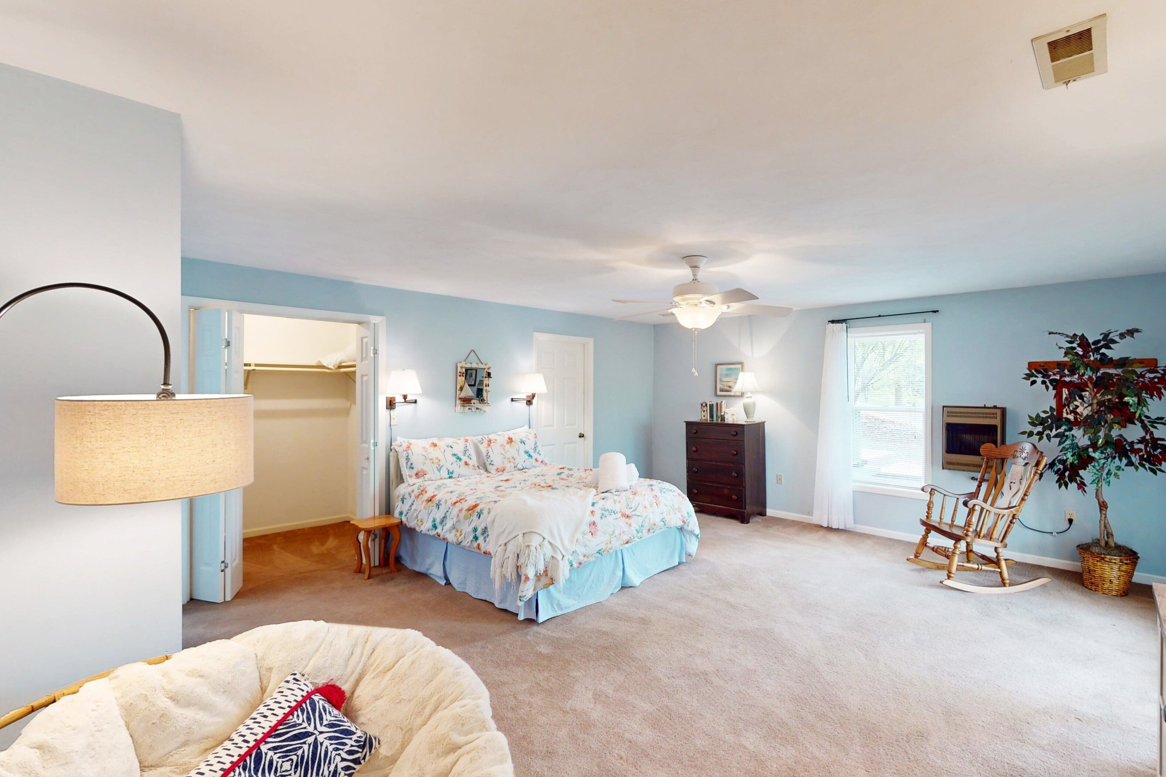 A bedroom with a bed, bedside lamps, a ceiling fan, a rocking chair, and a potted plant.