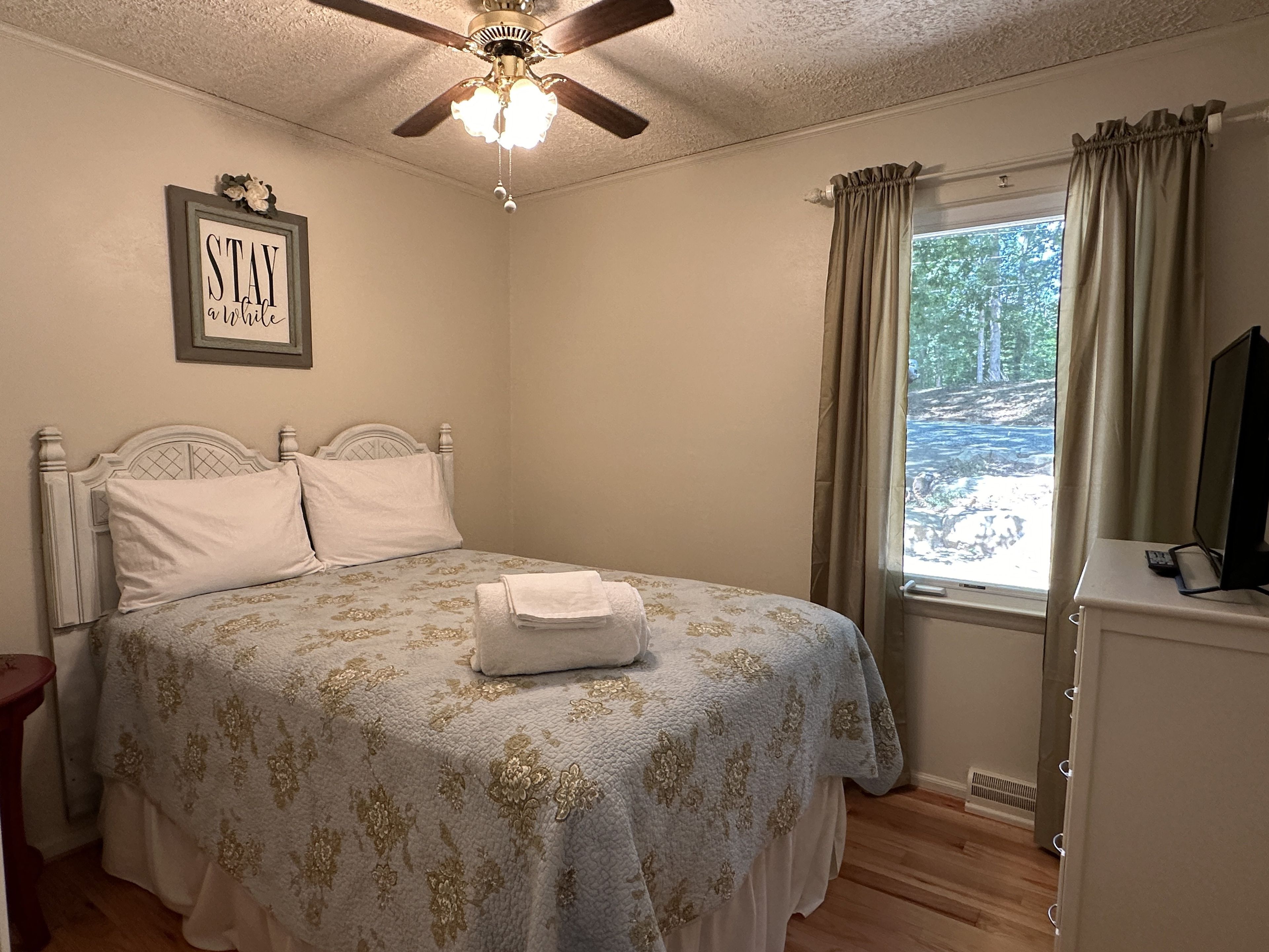 A bedroom with a bed, a ceiling fan, a window with curtains, and a bedside table.