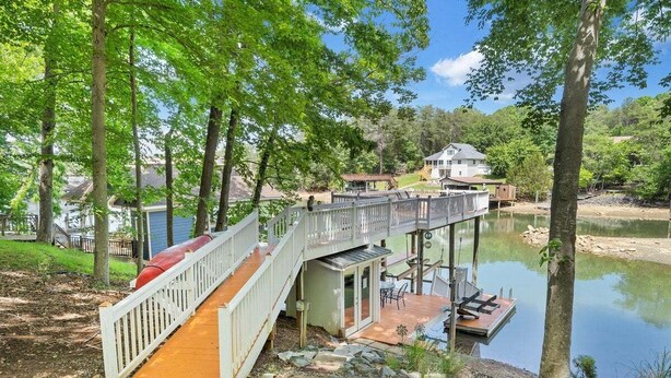 A wooden deck with a glass door leading to a covered outdoor area, a dock extending into a lake, and a house in the background. - Lakefront Tranquility - Lakefront Home With Views, Private hot Tub/dock, Deck, Free Wifi (Moneta)