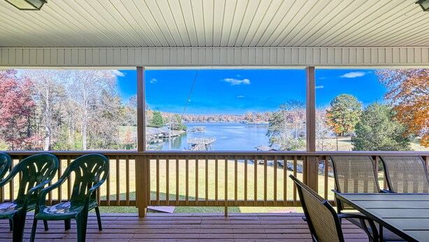A covered outdoor area with a view of a lake and trees, featuring a wooden deck, chairs, and a table. - Three Sheets to the Wind - Dog-friendly Lakefront Home With Dock, & Kayaks (Moneta)
