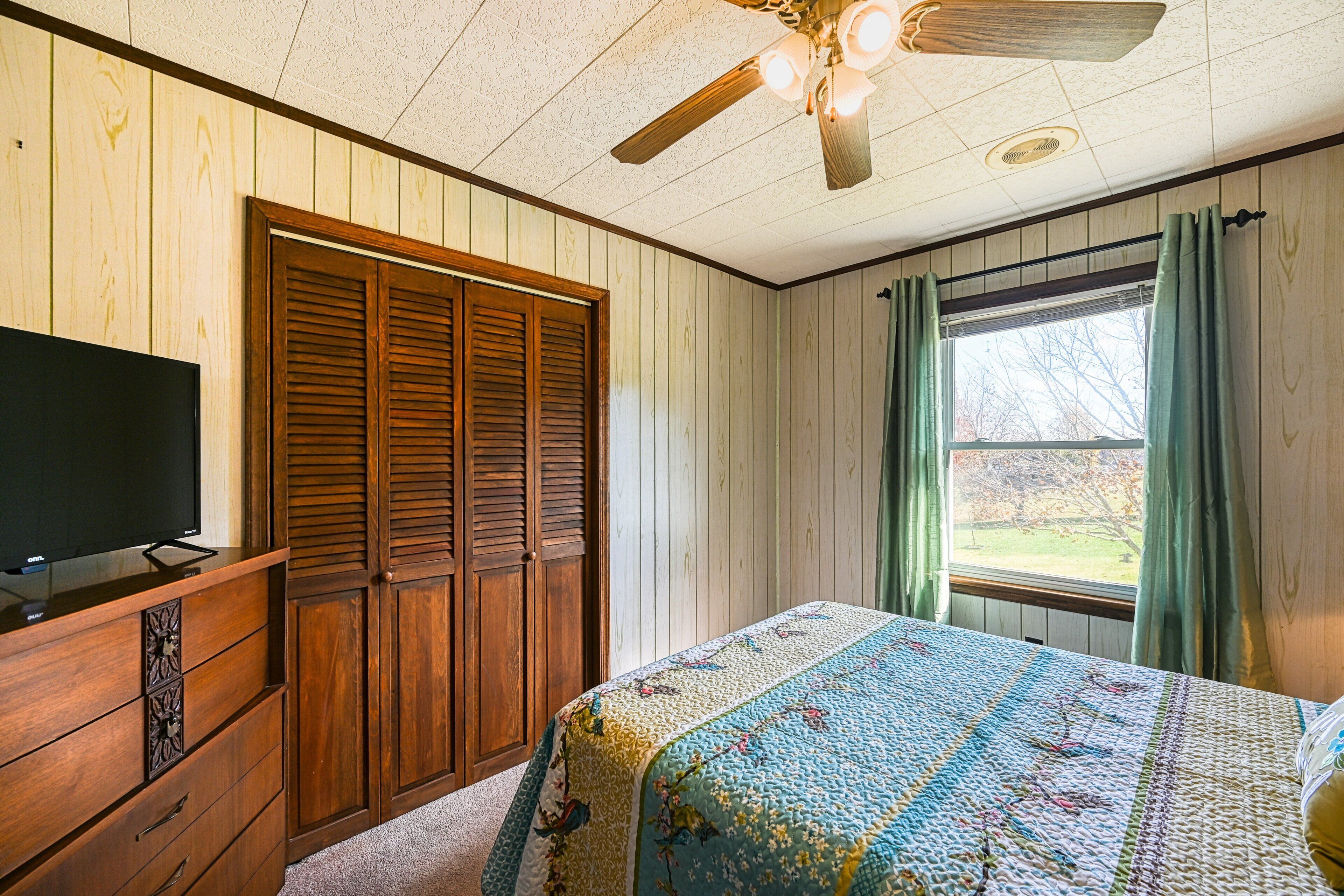 A bedroom with a wooden wardrobe, a bed with a quilt, a ceiling fan, and a window with curtains.