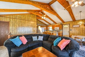 A cozy living room with a sectional sofa, wooden beams, and a kitchen in the background. - Three Sheets to the Wind - Dog-friendly Lakefront Home With Dock, & Kayaks (Moneta)