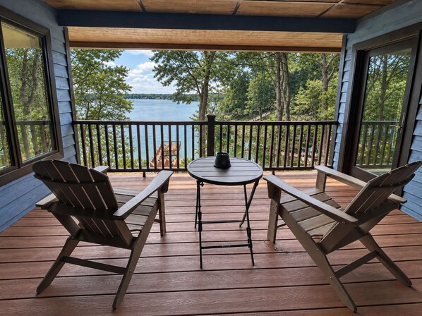 A wooden deck with two Adirondack chairs and a small round table, overlooking a lake with trees and a clear sky. - Lakedaze - Spacious Dog-friendly Lakefront House W/dock, hot tub & Putting Green (Moneta)