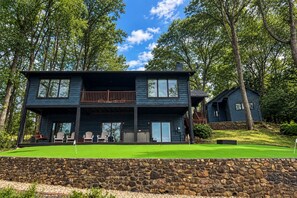 A two-story house with a balcony, a stone wall in the foreground, and a smaller building in the background. - Lakedaze - Spacious Dog-friendly Lakefront House W/dock, hot tub & Putting Green (Moneta)
