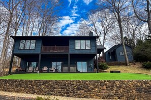 Une maison à deux étages avec un mur de soutènement en pierre, une pelouse et une terrasse avec des chaises.