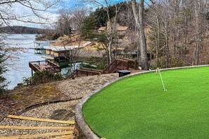 Un parcours de golf doté d’un green d’entraînement, d’une terrasse en bois et offrant une vue sur un lac et des bâtiments.