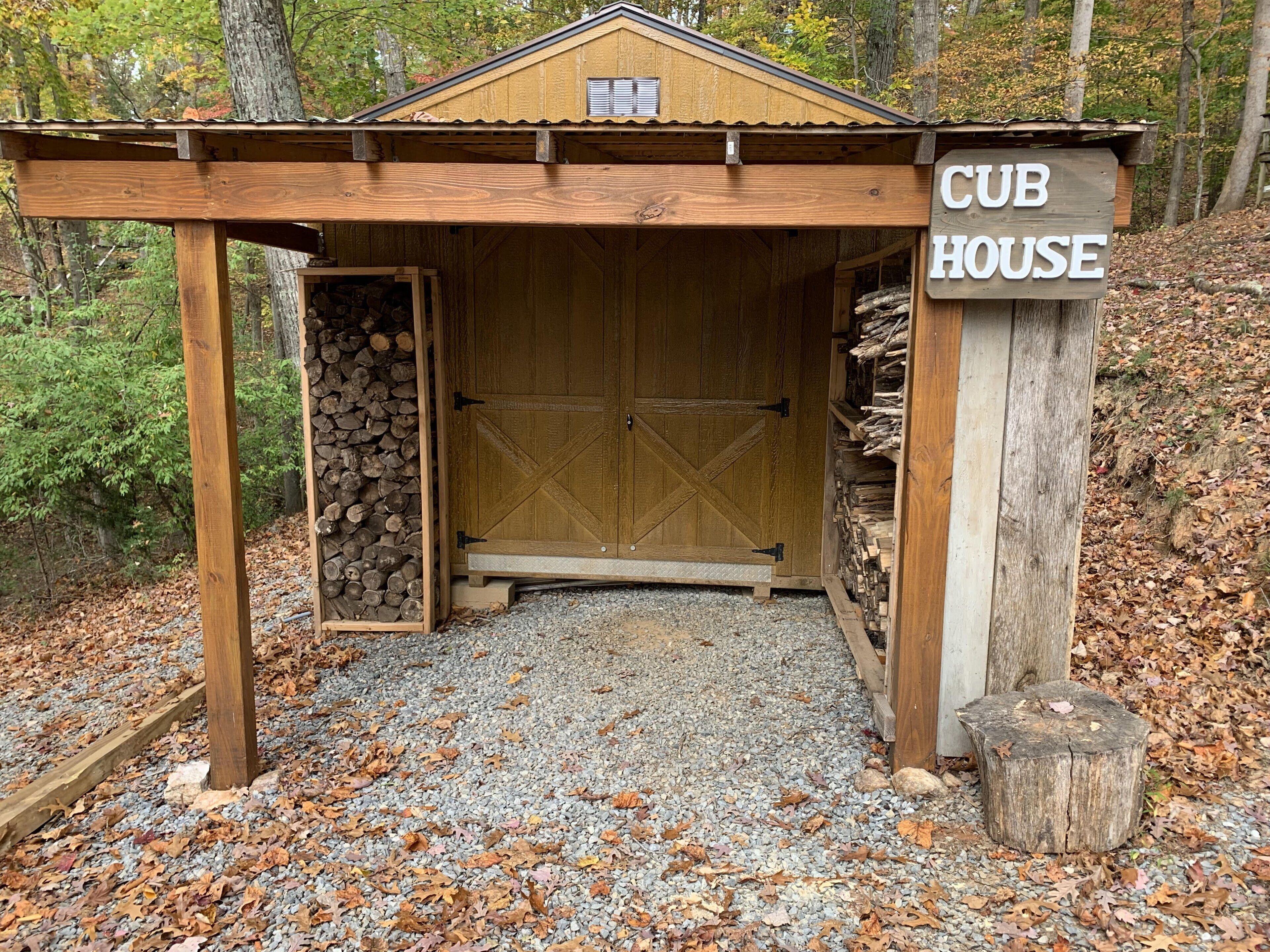 Una cabaña de madera con letrero 'CUB HOUSE', un camino de grava y una pila de leña afuera.